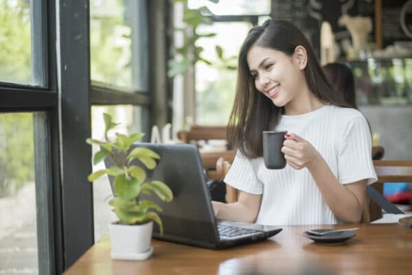 An asian woman drinking coffee from her mug while working on her laptop