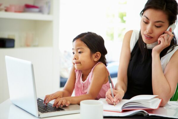 A busy mom talking on the phone and writing something down on a notebook while her child plays with her lapptop