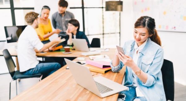 a young asian working using her phone in the workplace while her colleagues talk at the back