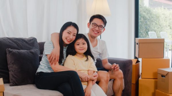 a happy young asian family sitting on a couch with cardboard boxes beside them