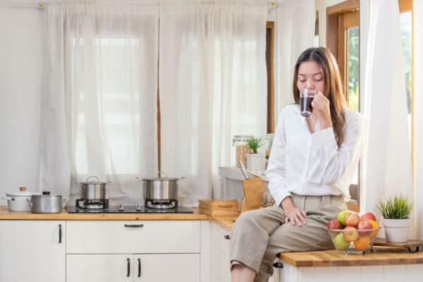 young beautiful Asian woman sitting on kitchen counter and drinking a cup of black coffee