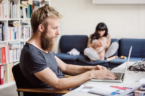 a man busy working on his laptop while his wife is looking after their baby